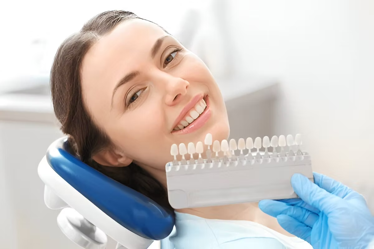 Woman in dental chair smiling while dentist holds tooth shade guide for implants